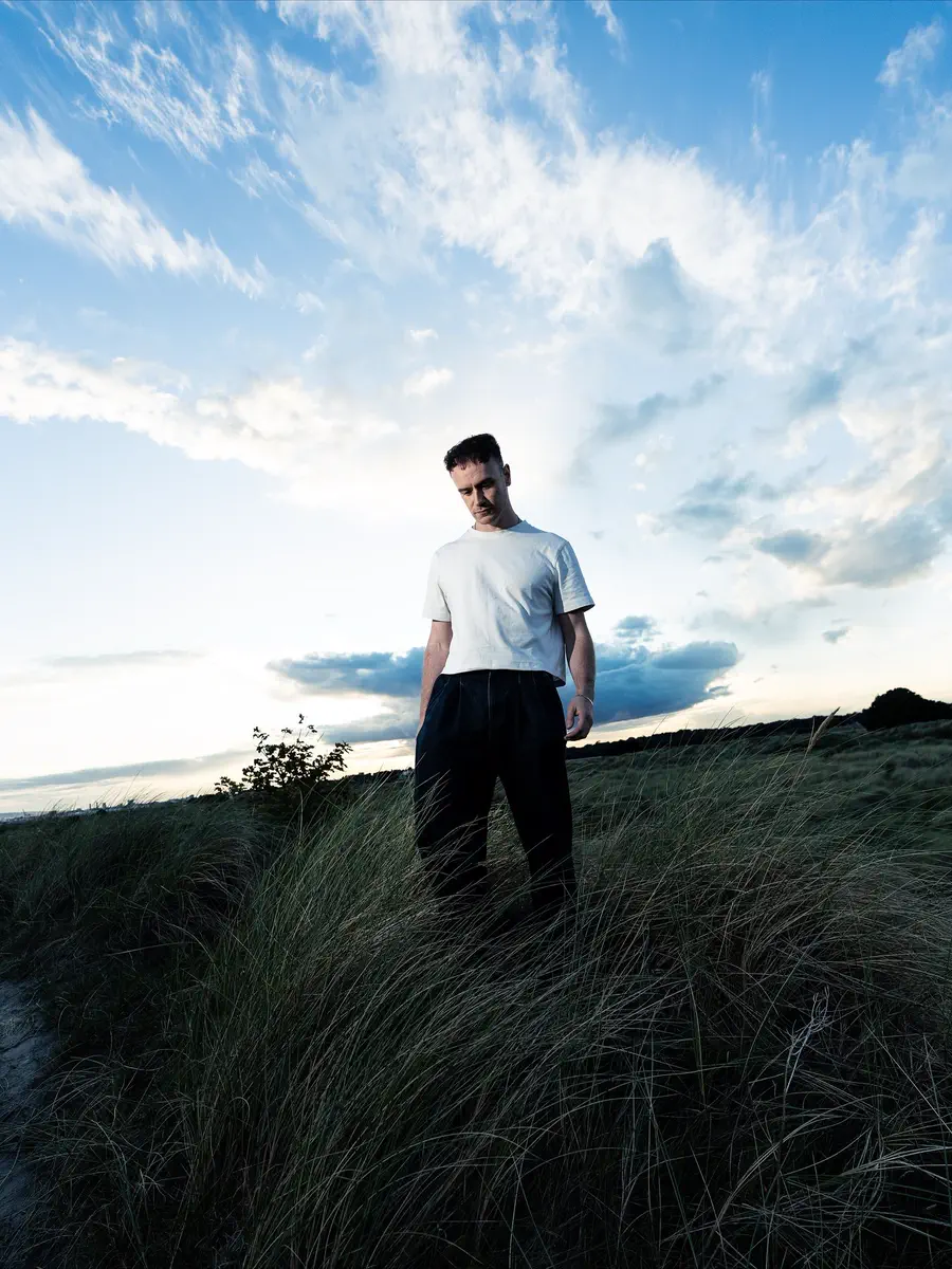Walshy standing in long dune grass under a wide evening sky, looking down